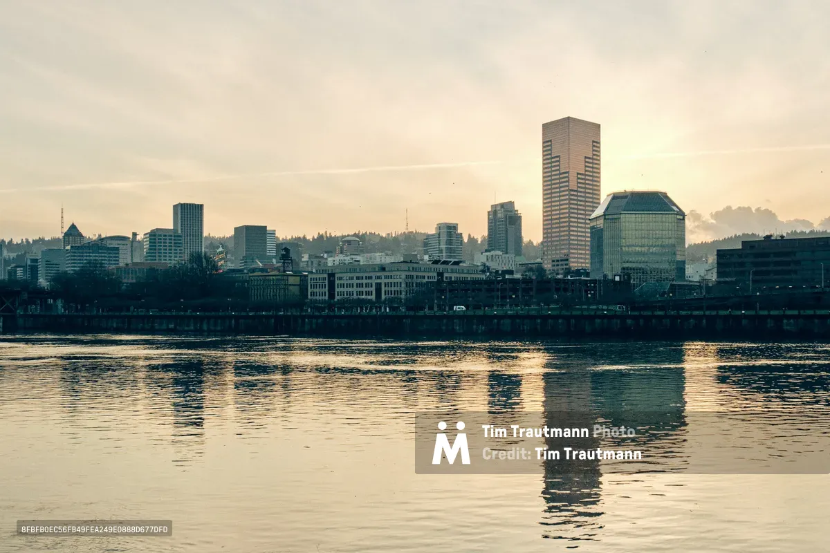 The Portland skyline emerges from forested hills in soft afternoon light, its modern towers and distinctive architecture casting perfect reflections across the rippling surface of the Willamette River. Shot from the Vera Katz Eastbank Esplanade, this composition captures the city's downtown core bathed in warm, golden hour luminosity with layered clouds stretching across the horizon. The water's gentle movement creates abstract patterns of light and shadow, while the silhouetted urban landscape transitions from glass and steel high-rises to the natural tree line beyond.