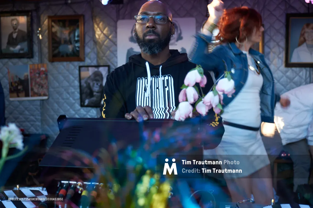 DJ Cee White commands the decks at White Owl Social Club during the season opener of Your Sunday Best day party in Portland, Oregon. Wearing glasses and a black hoodie with white text, he focuses intently on his laptop while a woman in motion blur dances behind him, pink flowers adding color to the intimate venue. The gallery walls adorned with framed artwork create an artistic backdrop for this vibrant Sunday afternoon gathering.