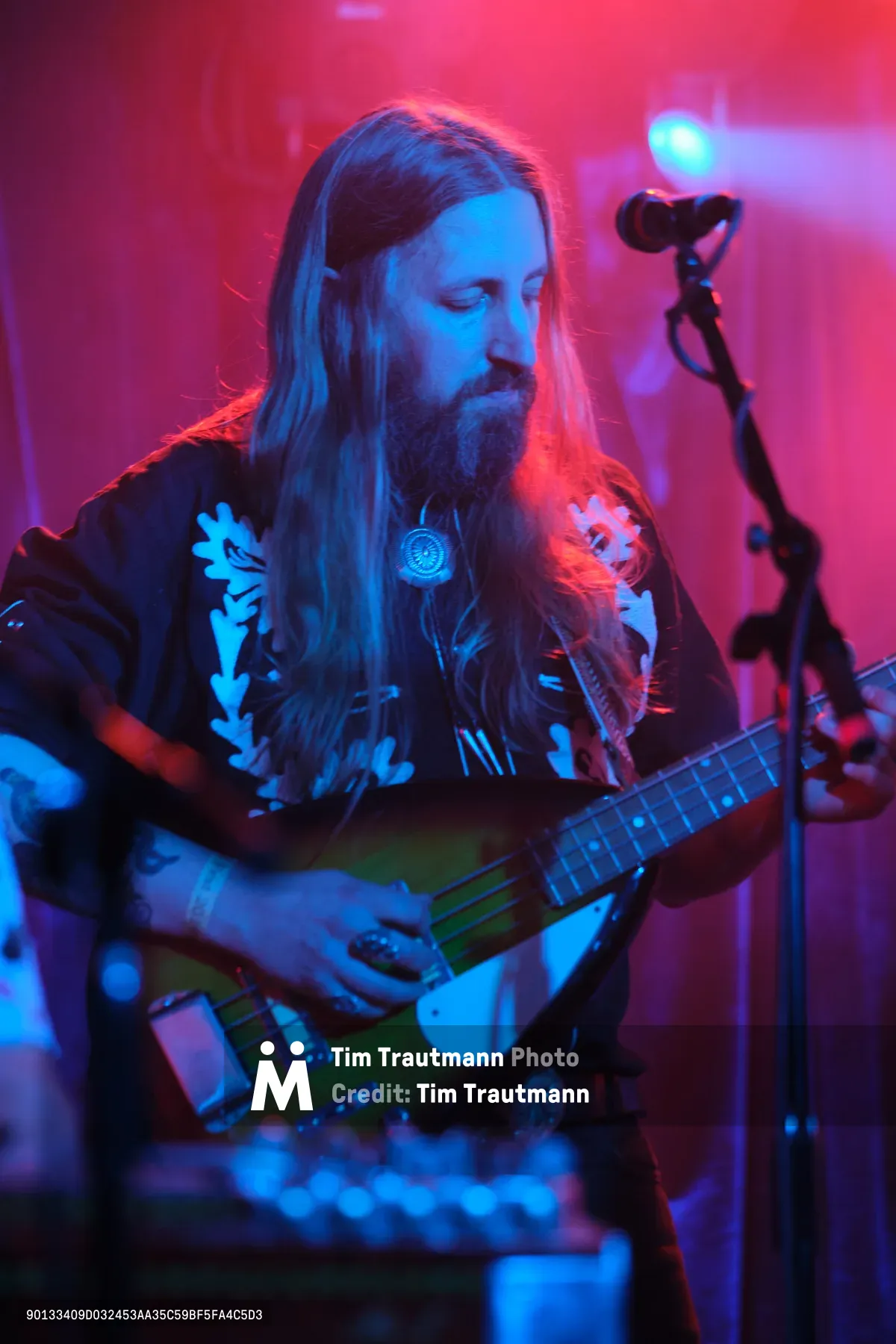 Tobias Berblinger of Roselit Bone commands the stage at Portland's legendary Dante's venue, his flowing blonde hair catching the saturated magenta and cyan stage lights. The bassist stands in contemplative focus, adorned in a black band tee and pendant necklace, while the atmospheric concert lighting transforms the intimate club into an otherworldly sonic cathedral. The image captures a moment of musical transcendence during RX Fest 2019, where underground music culture thrives in Portland's creative heart.