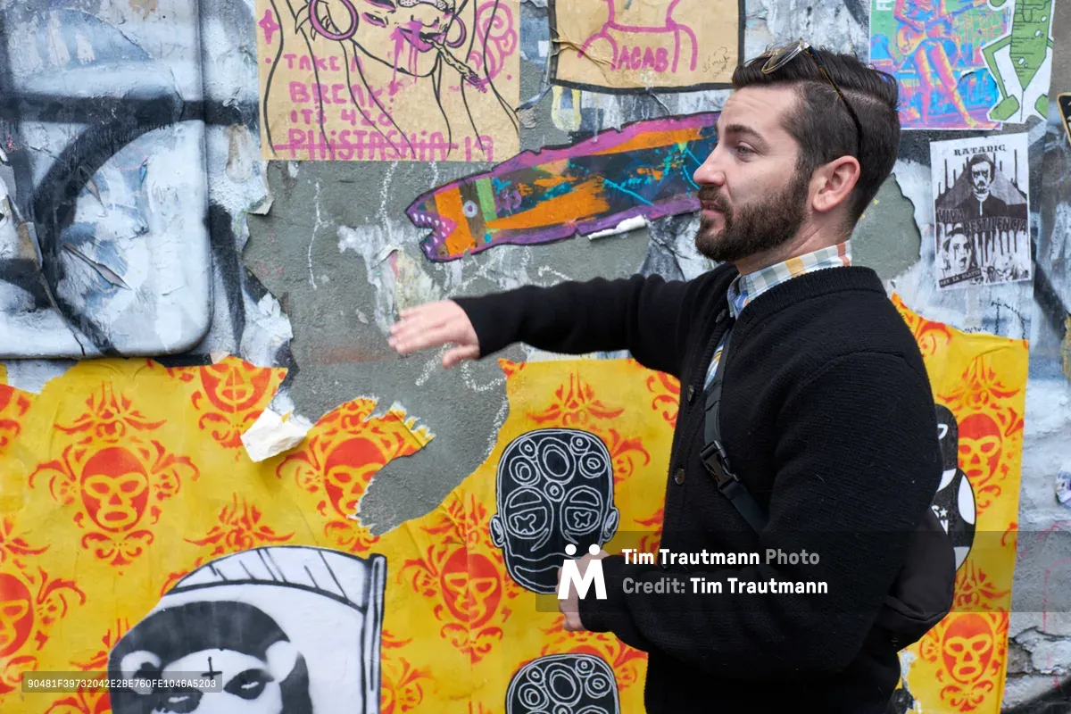 Tomás Valladares gestures enthusiastically toward a weathered concrete wall layered with vibrant street art, wheat paste installations, and graffiti in Portland's Central Eastside district. The bearded guide, dressed in a dark sweater, stands before a kaleidoscope of urban expression featuring yellow skull motifs, colorful paste-ups, and layered artwork that tells the story of the neighborhood's creative pulse. Behind him, the textured wall serves as a living gallery where artists have built upon each other's work, creating a palimpsest of contemporary street culture.