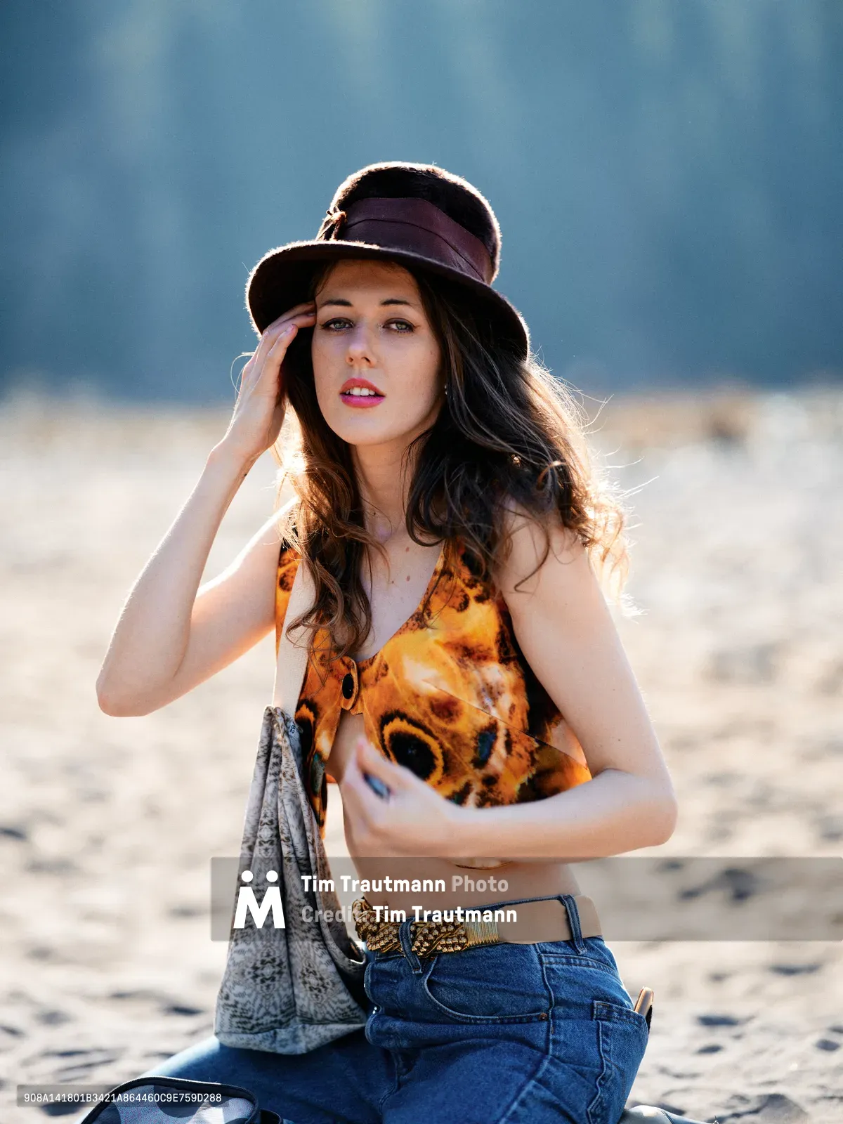 A young woman with flowing dark hair poses thoughtfully in the warm golden light of late afternoon along Oregon's Floodplain Trail in Multnomah County. She wears a burgundy wide-brimmed hat and vibrant leopard print top paired with high-waisted denim, her hand gracefully touching her temple in a contemplative gesture. The soft backlighting creates a luminous rim around her silhouette against the hazy blue-gray backdrop of the Columbia River Gorge landscape.