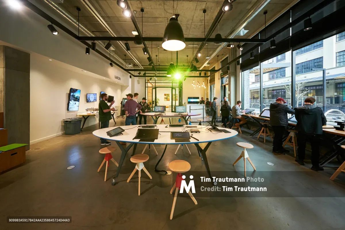 Inside the Wacom Experience Center in Portland's Pearl District, creative professionals gather around a striking circular white table equipped with digital tablets and laptops. The industrial-modern space features exposed ceiling infrastructure, pendant lighting, and floor-to-ceiling windows revealing the urban streetscape beyond. Colorful tripod stools in coral and natural wood surround the collaborative workspace, while additional visitors explore interactive displays and work stations throughout the open-plan facility.