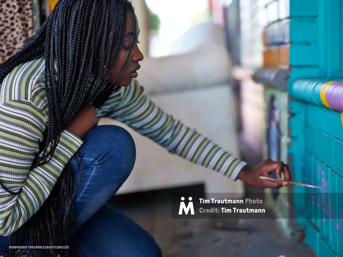 Naomi Likayi, wearing protective braids and a striped sweater, kneels beside her completed mural on Northeast 15th Avenue in Portland's Alberta Arts District. The afternoon light catches the intricate details of her box braids as she carefully signs her vibrant turquoise artwork, her concentrated expression reflecting the pride of artistic completion. The urban street setting showcases the neighborhood's commitment to public art and community expression.
