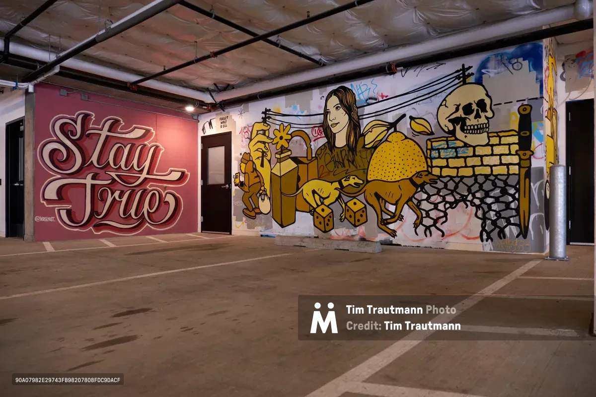 Vibrant street art transforms a concrete parking garage in Portland's Hosford-Abernethy neighborhood into an underground gallery. The commanding mural features bold lettering reading "Stay Girl" in deep burgundy alongside golden-hued figures including a woman with flowing hair, geometric shapes, and a skull motif against white walls. Industrial ceiling elements and marked parking spaces frame this juxtaposition of urban grit and artistic expression in the Taylor Electric Blocks.