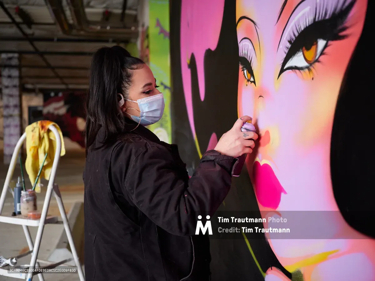Multidisciplinary artist Vilatose meticulously applies spray paint to a luminous female portrait mural within the industrial confines of Clay Creative's parking garage in Portland's Hosford-Abernethy neighborhood. Wearing a protective mask and dark hoodie, the artist works with focused precision on the dramatic face featuring amber eyes and flowing black hair rendered in electric pinks, purples, and coral hues. The raw concrete space transforms into an urban canvas, with paint supplies and a stepladder nearby suggesting hours of dedicated creative labor.