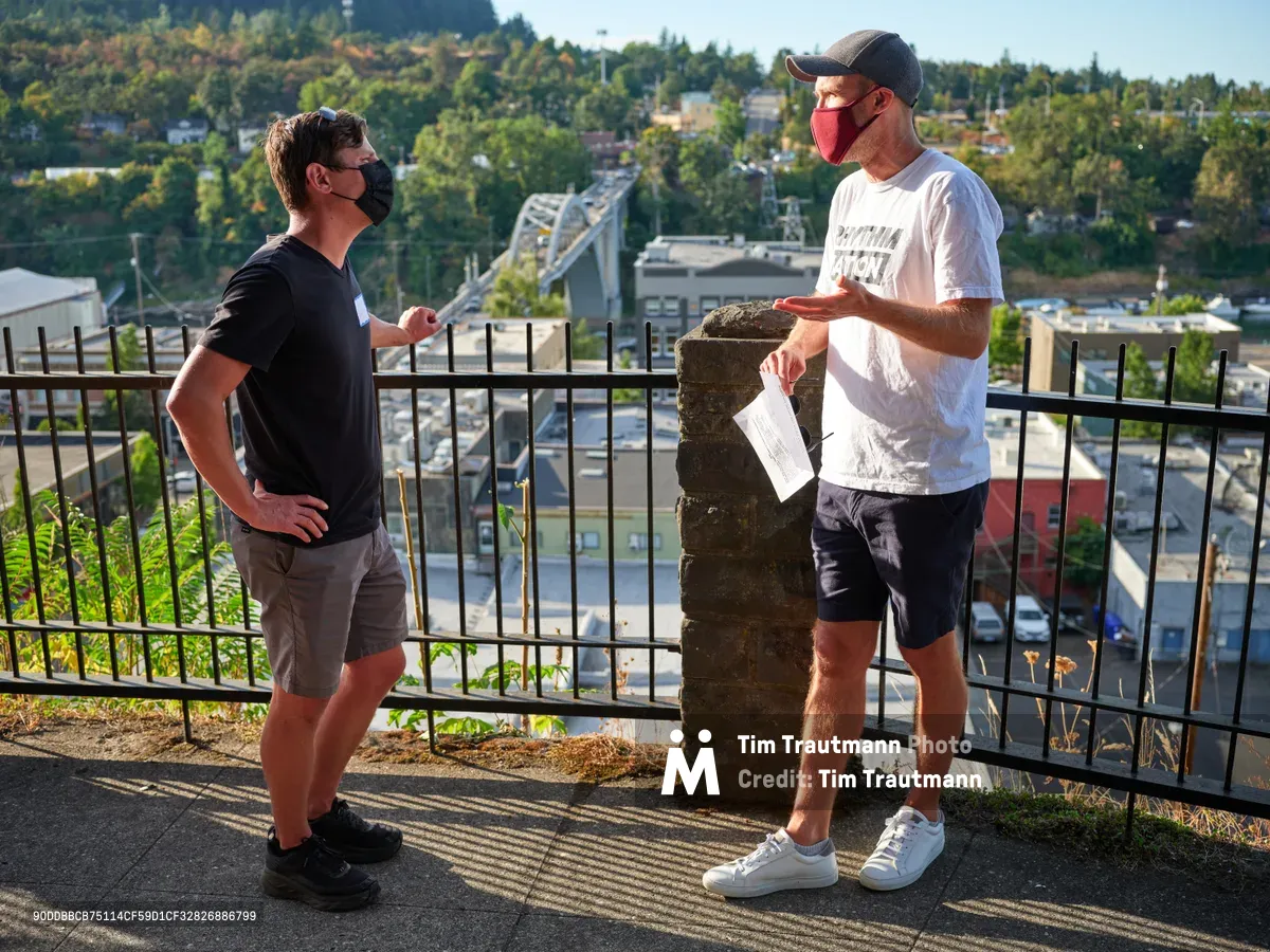 Michael Gaskill and Peter Marks engage in earnest conversation during a climate change protest at Representative Kurt Schrader's office in Oregon City. Standing against black iron railings with the Willamette River valley and forested hills stretching behind them, both men wear face masks in the golden afternoon light. The intimate moment captures the grassroots nature of political activism, with Marks holding papers while gesturing toward his companion against the backdrop of the McLoughlin Promenade overlook.
