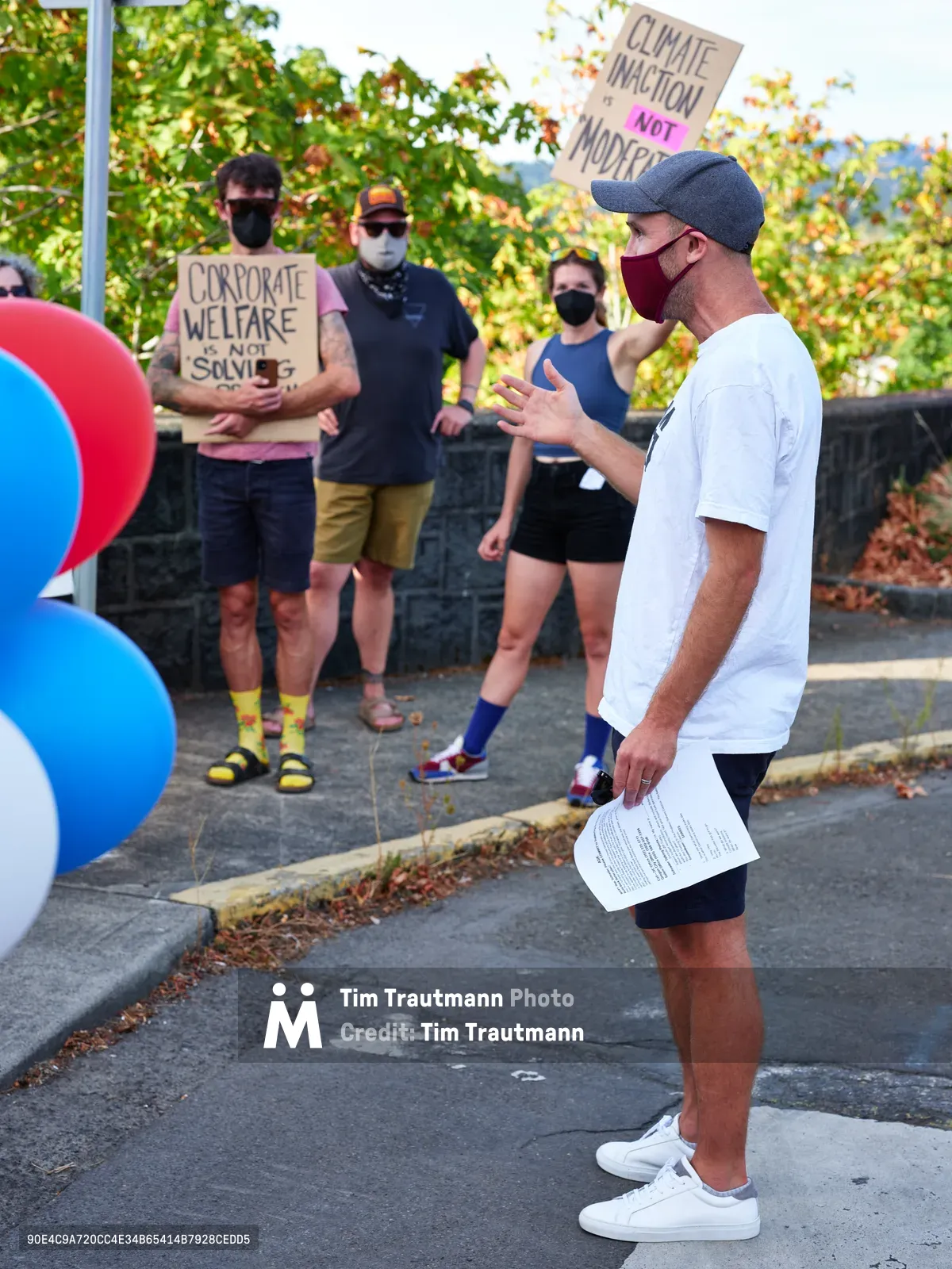 Organizer Peter Marks gestures emphatically while speaking to a small crowd of masked protesters gathered outside Representative Kurt Schrader's office in Oregon City. The speaker, wearing a white t-shirt and baseball cap, holds papers while addressing activists holding signs reading "Climate Inaction Not Modest" and "Corporate Welfare is Not Science." Autumn foliage creates a warm backdrop against the urban setting of McLoughlin Promenade, capturing a moment of grassroots political engagement during the pandemic era.