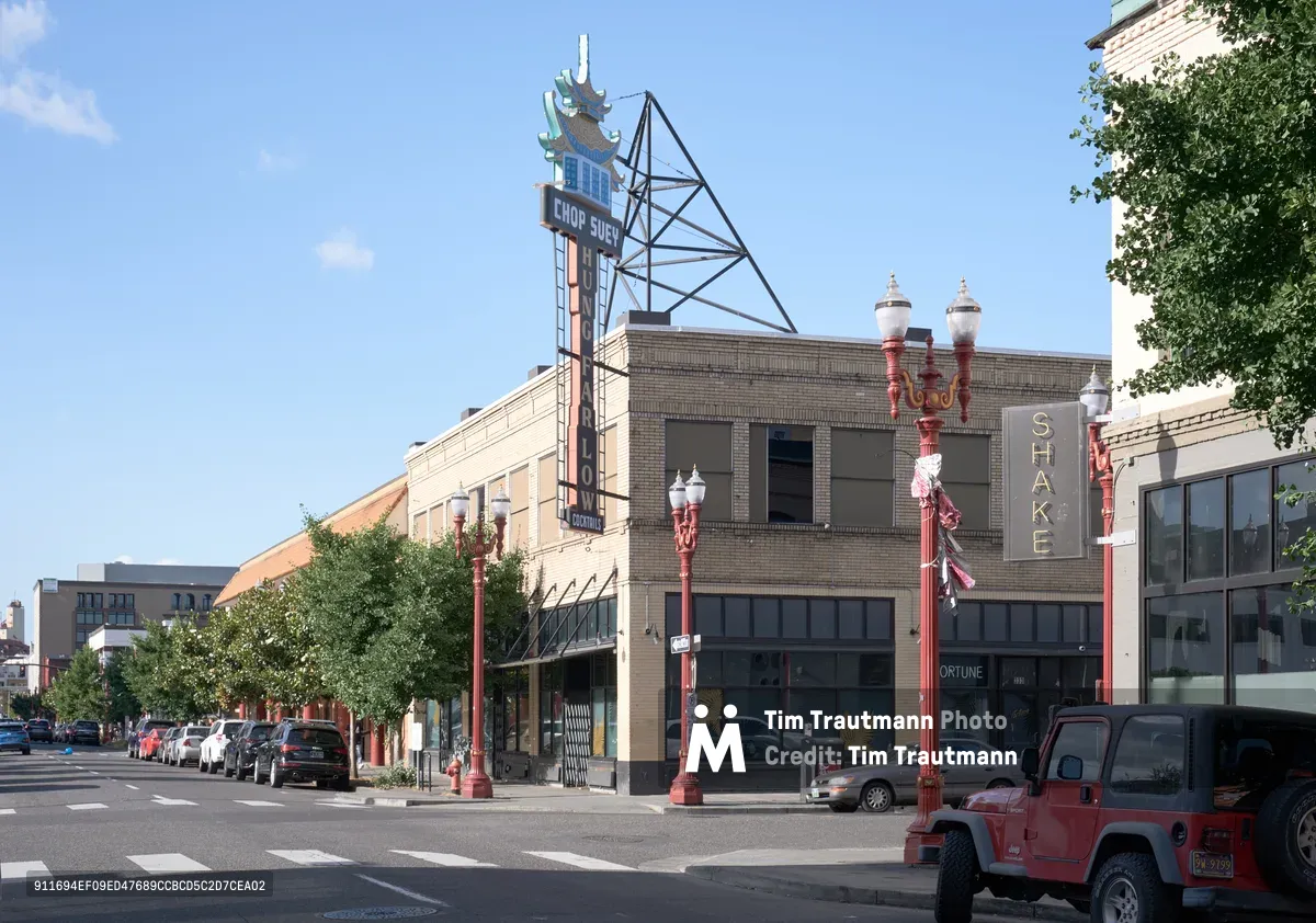A vintage neon sign for 'Chop Suey' crowns a brick building along a tree-lined street in Portland's historic Chinatown district. The iconic pagoda-topped sign, supported by industrial scaffolding, stands as a cultural landmark against a clear blue afternoon sky. Red ornate street lamps line the avenue while parked cars and storefronts including 'Shake' create a tableau of urban heritage preservation.