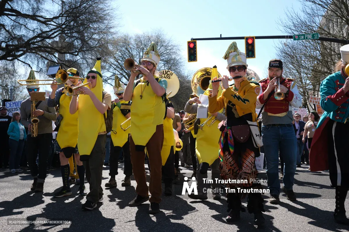 A spirited brass band dressed in bright yellow costumes and elaborate crowns commands the intersection at Southwest Pine Street in Portland's Old Town district during the March 2026 No Kings protest. The musicians, calling themselves the Unpresidented Brass Band, blow trumpets and tubas while marching through bare winter trees under crisp blue skies. Their theatrical performance transforms political dissent into carnival-like spectacle, drawing crowds of onlookers who gather beneath the traffic lights at this historic downtown crossroads.