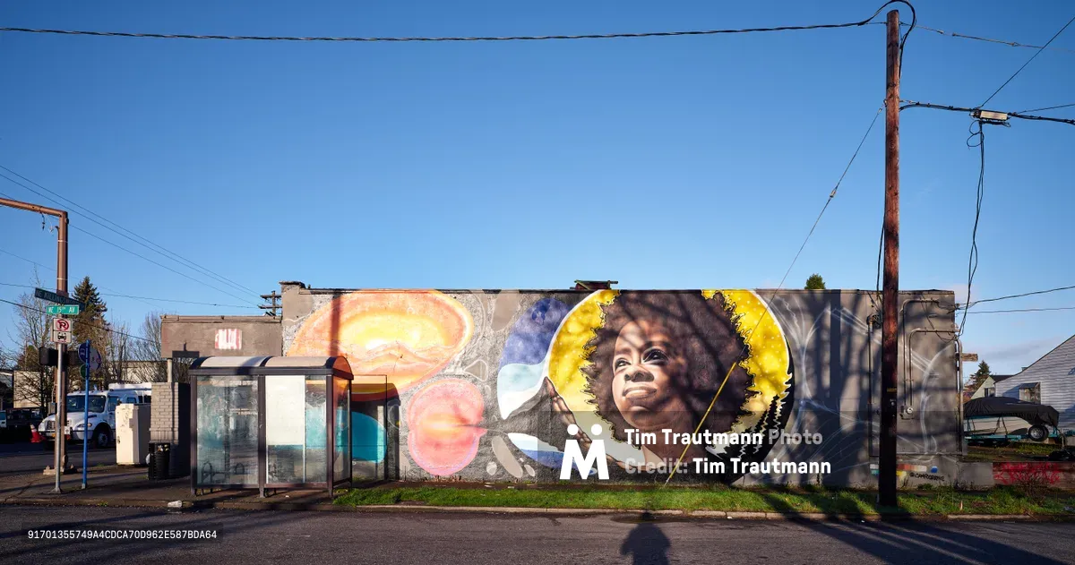A vibrant street mural depicting poet Amanda Gorman adorns the side wall of a commercial building at the intersection of Northeast 15th and Killingsworth in Portland's Alberta district. The artwork features Gorman's portrait surrounded by golden celestial imagery and cosmic elements, rendered in warm yellows and cool blues against the clear winter sky. Power lines crisscross overhead while bare tree branches frame the scene, creating an urban intersection where art transforms everyday infrastructure into a celebration of literary achievement.