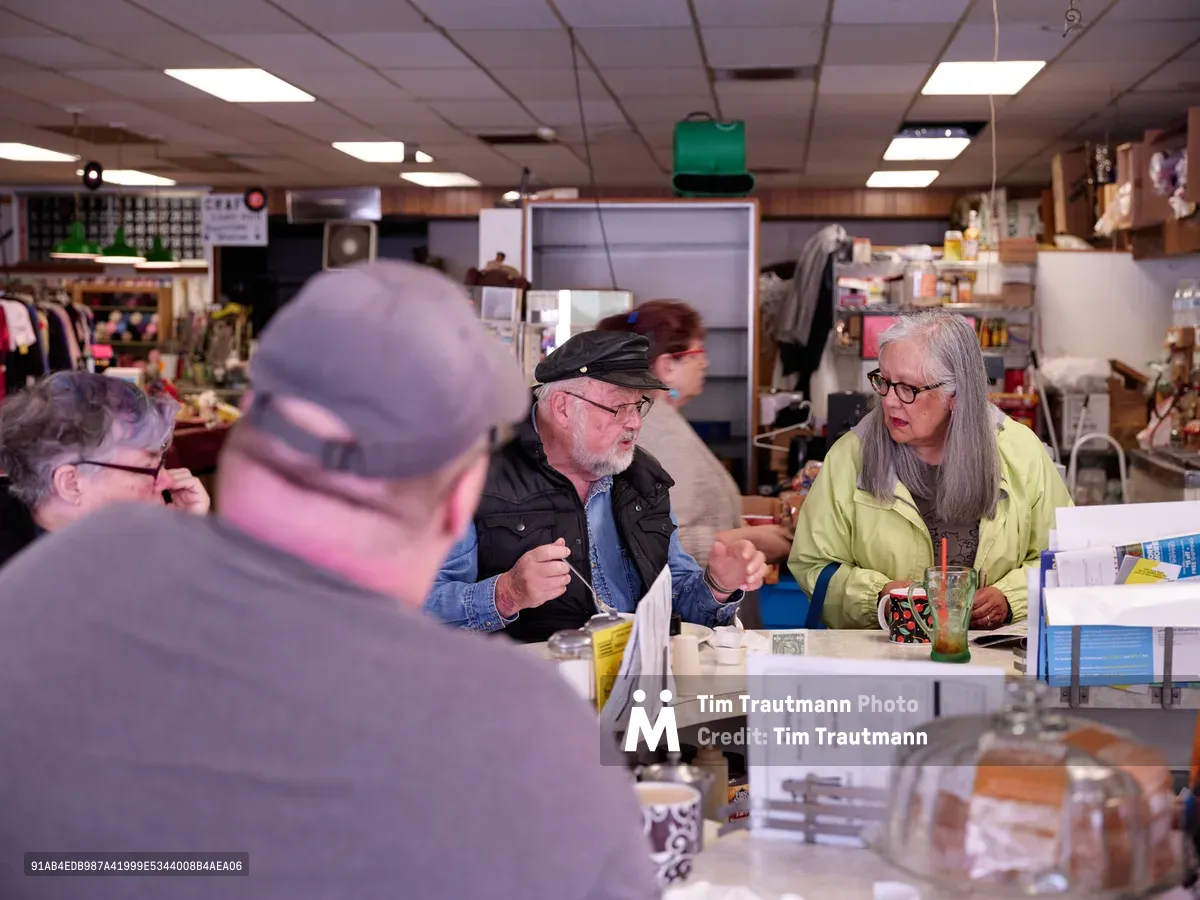 Under the warm glow of fluorescent ceiling lights, patrons gather at the worn formica counter of Pattie's Home Plate Café in Portland's Saint Johns neighborhood. A bearded man in a black cap and vest leans forward in animated conversation with a woman in a bright yellow jacket, her long gray hair catching the light as she gestures expressively. The intimate counter scene captures the timeless ritual of neighborhood dining, where scattered newspapers, coffee cups, and the cluttered backdrop of a working kitchen create an authentic portrait of blue-collar community life.