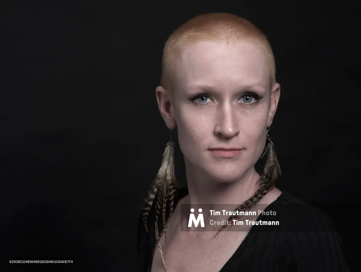 Cayce Ryan gazes directly at the camera with quiet intensity, her closely-cropped hair catching the controlled studio lighting against the deep black backdrop. Feathered earrings frame her face, adding organic texture to the minimalist composition, while her subtle makeup and black garment create an understated elegance. The professional headshot, captured in Portland's Factor Building, reveals both vulnerability and strength through precise lighting that sculpts her features with dramatic shadow and highlight.