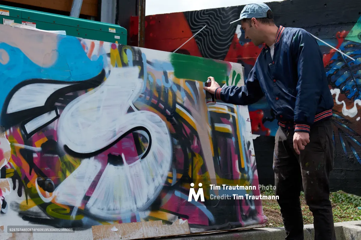 Galen Malcolm of the Portland Street Art Alliance crouches beside a vibrant mural in progress, spray can in hand, during a live demonstration at the Taylor Electric Blocks in Portland's Hosford-Abernethy neighborhood. The artist wears a gray baseball cap and navy jacket with red trim, his focused posture capturing the concentrated energy of urban art creation. Behind him, layers of colorful graffiti cover concrete walls in explosive bursts of blue, white, pink, and yellow, creating a kaleidoscopic backdrop that speaks to Portland's thriving street art culture. The scene unfolds in the industrial-artistic environment of Clay Creative on Southeast 3rd Avenue, where raw concrete meets creative expression.
