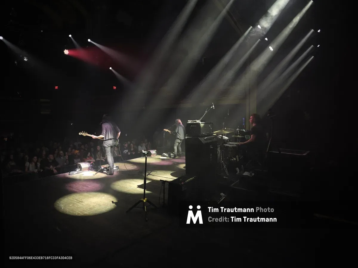 Emma Richardson, Julian Dorio, and Russell Marsden of Band of Skulls perform during their 10 Year Anniversary Tour at Manhattan's historic Webster Hall. Dramatic stage lighting cuts through the venue's darkness, casting golden pools across the performance space while silhouetting the trio against towering light beams. The intimate concert setting captures the raw energy of live rock performance, with the audience gathered in shadow around the illuminated stage.