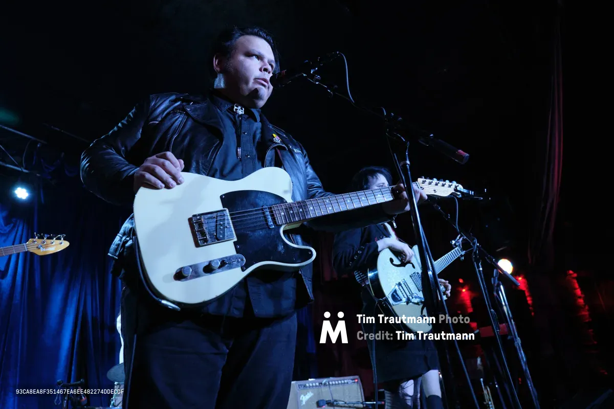 Victor Franco of Roselit Bone grips his white Telecaster guitar under dramatic blue stage lighting at Dante's in Portland, Oregon during RX Fest 2019. The leather-jacketed performer stands focused at the microphone, his intense gaze cutting through the moody concert atmosphere while bandmates remain partially visible in the shadowed background. The intimate venue's low ceiling and close quarters create an electric tension between artist and audience.