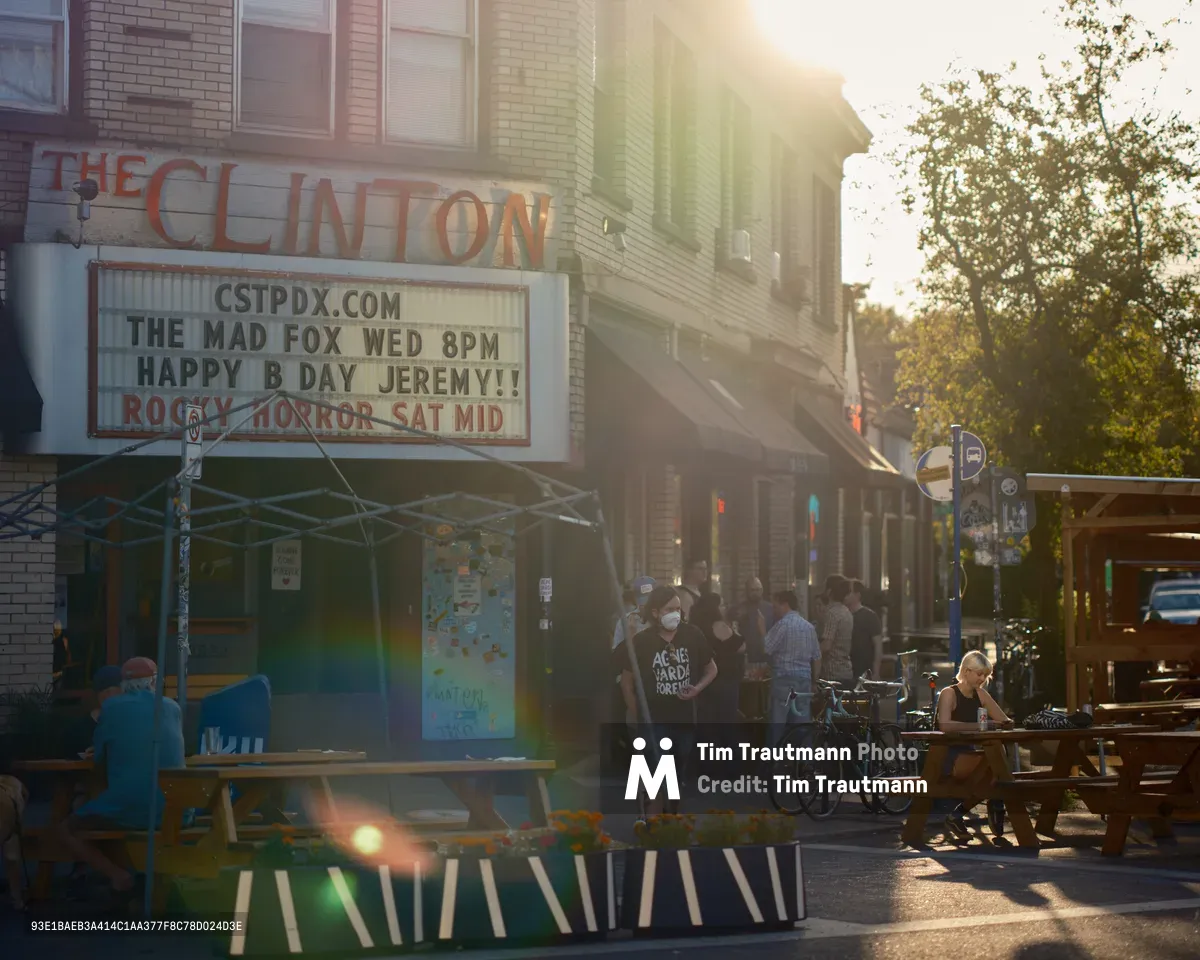 The exterior of The Clinton Street Theater in Portland, Oregon, bathed in warm golden lens-flare sunlight. The theater's marquee reads "The Mad Fox Wed 8PM — Happy B Day Jeremy!! — Rocky Horror Sat Mid." A lively crowd of people gathers on the sidewalk outside, with outdoor picnic tables and planters with marigolds occupying a parklet in the foreground. A woman in a face mask wearing a black "Agnès Varda Forever" t-shirt walks past. Bicycles are parked along the curb, and a woman sits alone at a picnic table to the right.