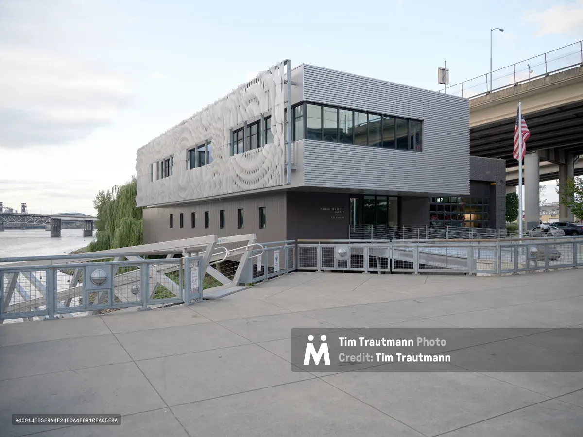 Portland Fire & Rescue Station 21 presents a striking contemporary facade beneath the evening's pewter sky, its sculptural white upper level contrasting sharply with the utilitarian concrete base. The building's distinctive undulating exterior panels create dramatic shadows while large glass windows reveal the operational spaces within. An American flag stands sentinel beside the waterfront structure, with the Willamette River and distant bridges completing this scene of municipal modernism.