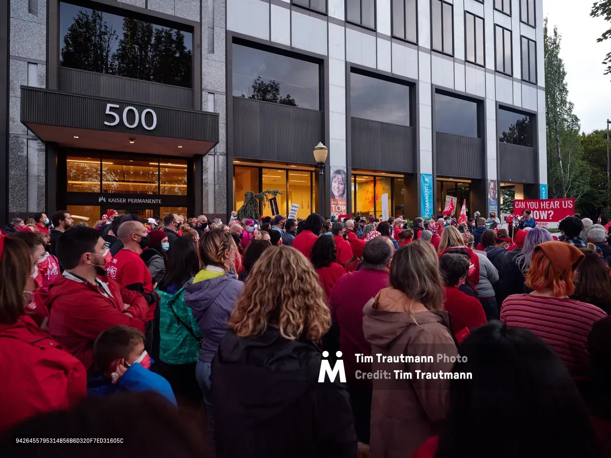 A sea of red-clad healthcare workers fills the sidewalk outside Kaiser Permanente's modernist headquarters at 500 Northeast Multnomah Street in Portland's Lloyd District. The crowd of striking nurses creates a powerful visual contrast against the building's sleek glass facade and concrete columns, their unified presence illuminated by the warm glow spilling from the medical center's ground-floor windows in the gathering dusk. Signs demanding "CAN YOU HEAR US NOW?" punctuate the peaceful but determined assembly, while the institutional architecture looms overhead, emphasizing the David-versus-Goliath nature of the labor dispute.