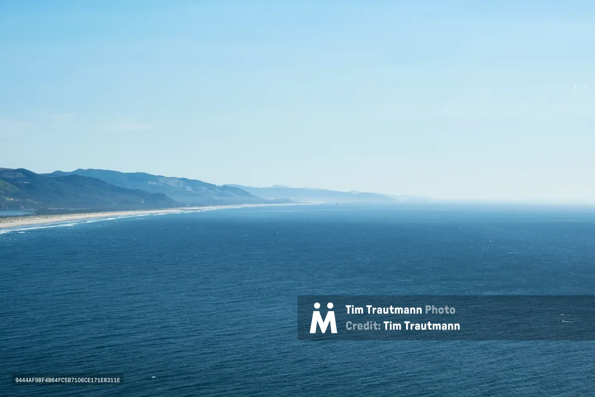 An expansive aerial view captures the Oregon coast where the Pacific Ocean meets the rugged Tillamook County shoreline. The deep blue waters stretch endlessly toward a hazy horizon, while forested mountains roll inland from a pristine sandy beach. Soft atmospheric haze creates layers of blue tones that blend sea, sky, and distant peaks into a seamless coastal panorama.