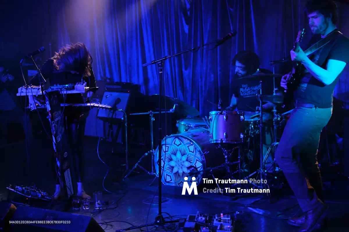 Sun Blood Stories delivers an atmospheric performance bathed in ethereal blue and purple stage lighting at Dante's in downtown Portland. The trio creates an intimate soundscape with synthesizers, drums, and guitar, their silhouettes emerging from the moody wash of color against draped curtains. The geometric mandala design on the drum kit serves as a focal point amid the sea of electronic equipment and cables sprawling across the venue floor.