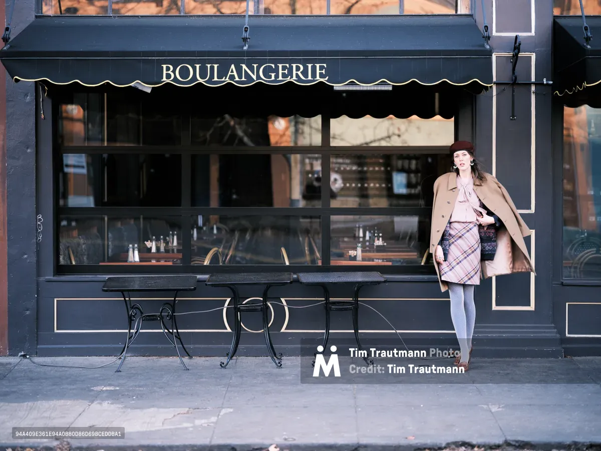A woman in autumn layers—camel coat, plaid skirt, and burgundy beret—poses beside a charming French boulangerie on Northeast Alberta Street in Portland, Oregon. The dark navy storefront with its classic awning and wrought iron café tables creates an intimate European atmosphere against the urban Pacific Northwest backdrop. Golden hour light softly illuminates the scene, casting warm reflections through the café's large windows while highlighting the contrast between the woman's vintage-inspired ensemble and the contemporary street setting.