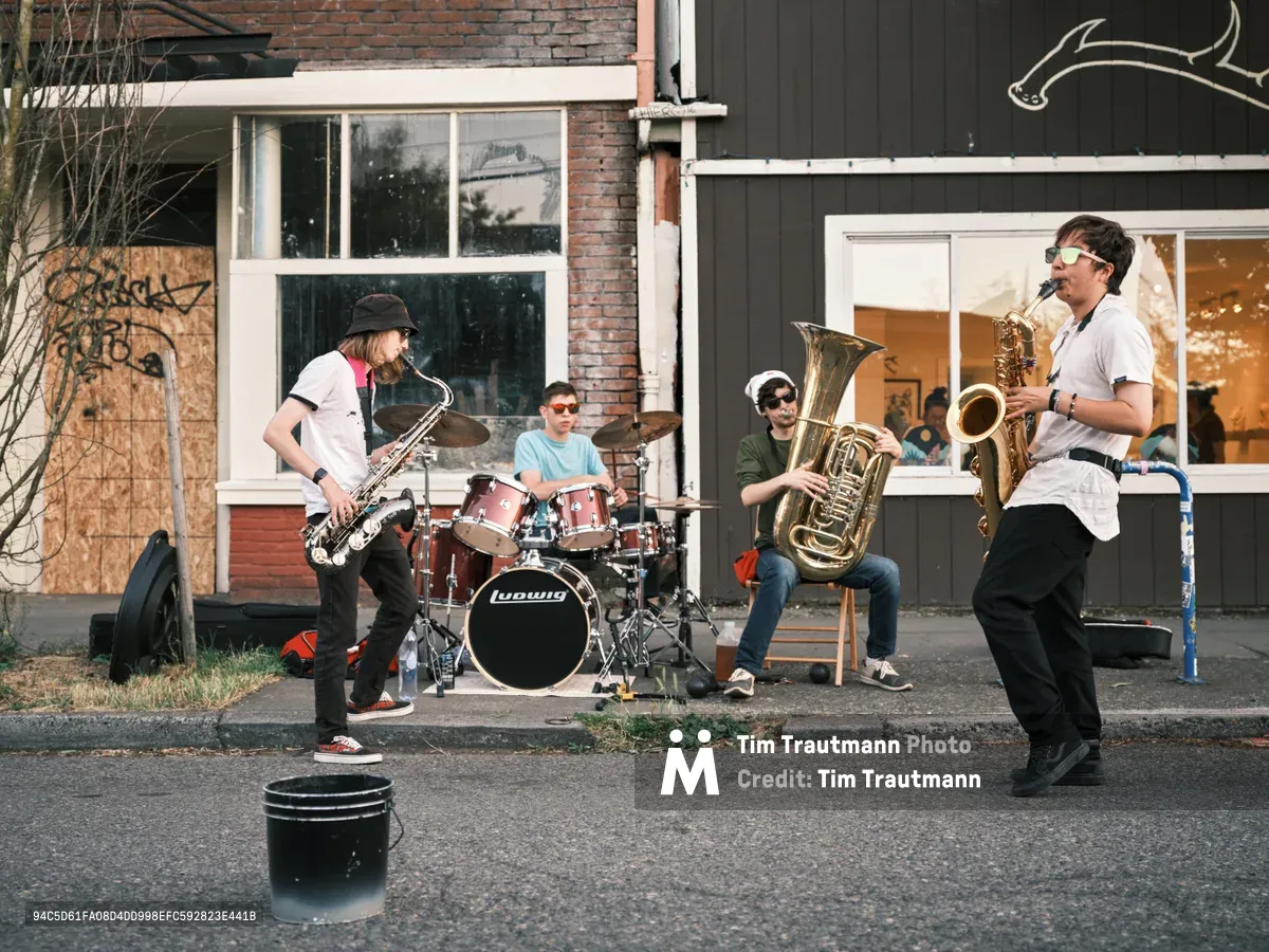 Four young musicians perform an impromptu jazz session on the weathered asphalt of Northeast Alberta Street during Portland's Last Thursday art walk. Golden afternoon light bathes the scene as a saxophonist in a black beanie leads the ensemble, while a drummer, tuba player, and second saxophonist create an intimate street corner concert. Behind them, the characteristic mix of brick and dark wood siding reflects the gentrifying neighborhood's evolving character, with curious onlookers visible through storefront windows.