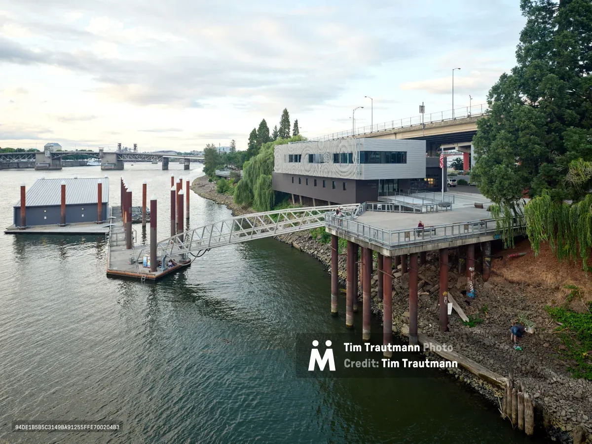 Portland Fire & Rescue Station 21 emerges from the Willamette River shoreline like a modernist beacon, its angular concrete form cantilevering over the water on weathered steel pilings. The facility's distinctive floating dock system extends into the jade-green current, while the Southeast Madison Street bridge spans the horizon under a brooding Pacific Northwest sky. This architectural marvel represents the intersection of urban emergency services and riverfront infrastructure, captured during the soft light of an overcast afternoon.