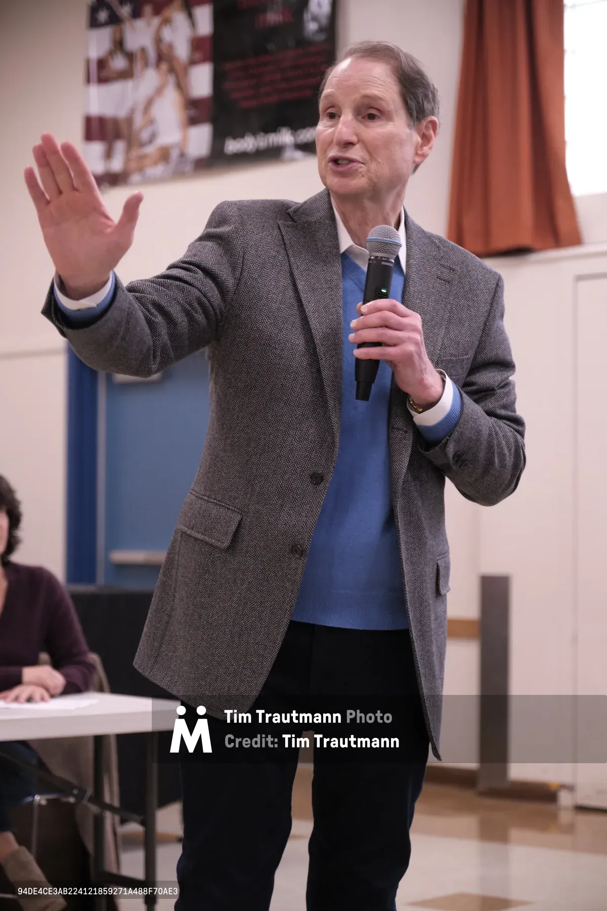 Senator Ron Wyden gestures emphatically while speaking into a wireless microphone during a town hall meeting at Robert Gray Middle School in Portland's Hillsdale neighborhood. The Democratic senator, wearing a textured gray blazer over a blue sweater, raises his left hand while addressing constituents in the school's multipurpose room. Patriotic posters and educational materials create a backdrop of civic engagement in this intimate community gathering.