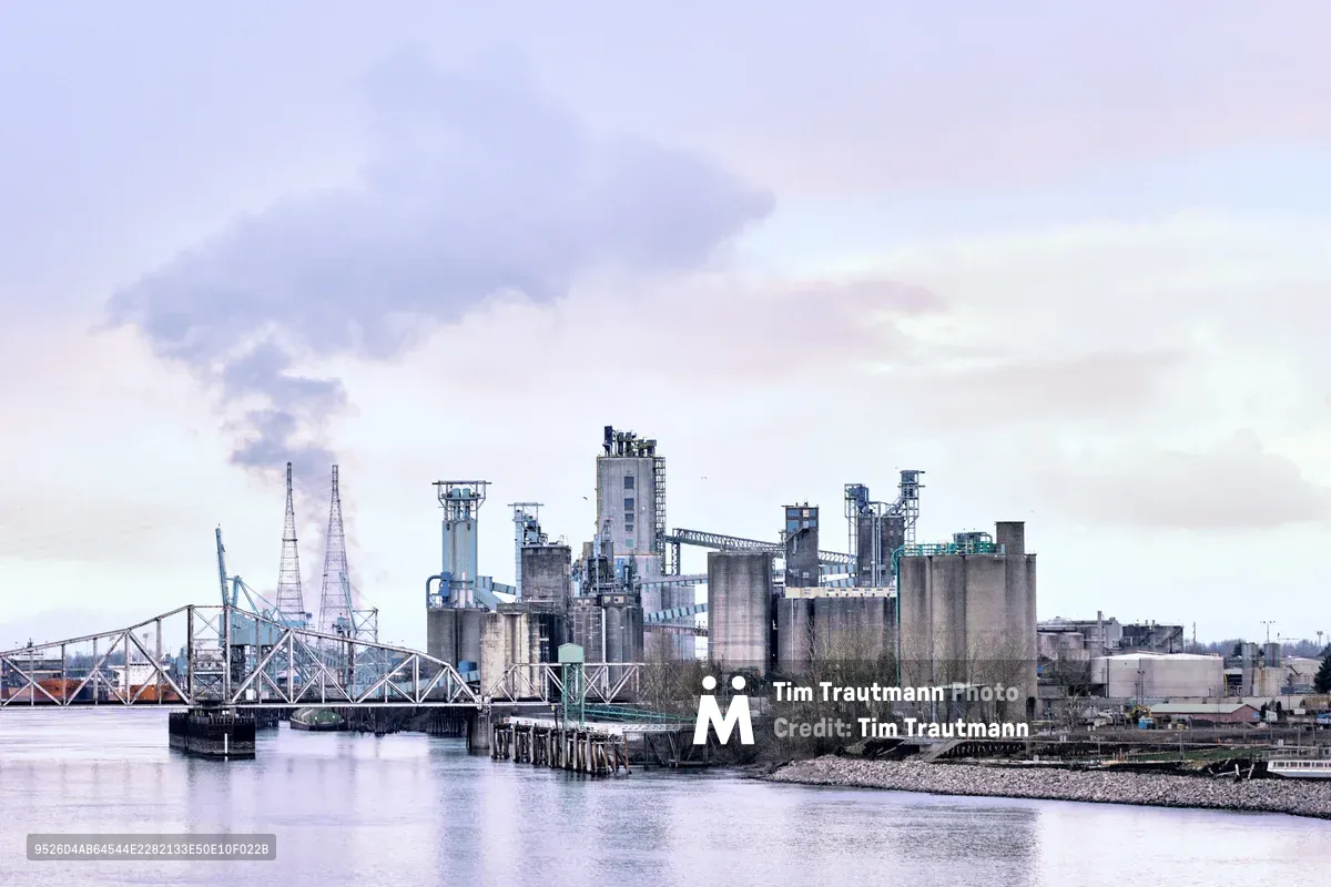 The United Grain Corporation's Vancouver Export Terminal rises from the Columbia River's edge in a symphony of concrete silos, steel conveyor bridges, and industrial towers. Captured during the warm afternoon hours, the facility's imposing structures are bathed in soft lavender and rose-tinted light, their reflections shimmering across the glassy water. A steel truss bridge spans the river in the left frame, connecting this vital grain export hub to the broader Pacific Northwest transportation network.