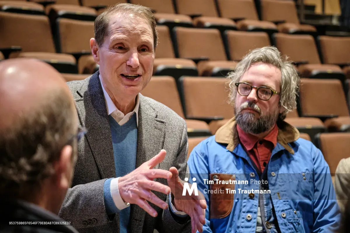 Senator Ron Wyden gestures expressively while speaking to constituents in a lecture hall setting in Portland's Saint Johns neighborhood. The Oregon Democrat, wearing a herringbone blazer over a blue sweater, commands attention with animated hand movements as he engages with community members. Behind him, a bearded constituent in a denim jacket listens intently, while tiered wooden seating creates an intimate academic atmosphere for this town hall gathering.