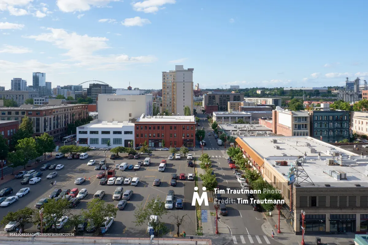 An elevated view captures Portland's historic Chinatown district on a bright afternoon, where brick facades and contemporary structures create an eclectic urban tapestry. The bustling street scene unfolds below with diagonal parking lots filled with vehicles, tree-lined sidewalks, and pedestrians navigating the intersection of heritage and modern commerce. Puffy white clouds drift across azure skies above this Northwest Portland neighborhood, while the distinctive architecture tells the story of cultural evolution in the Pacific Northwest.
