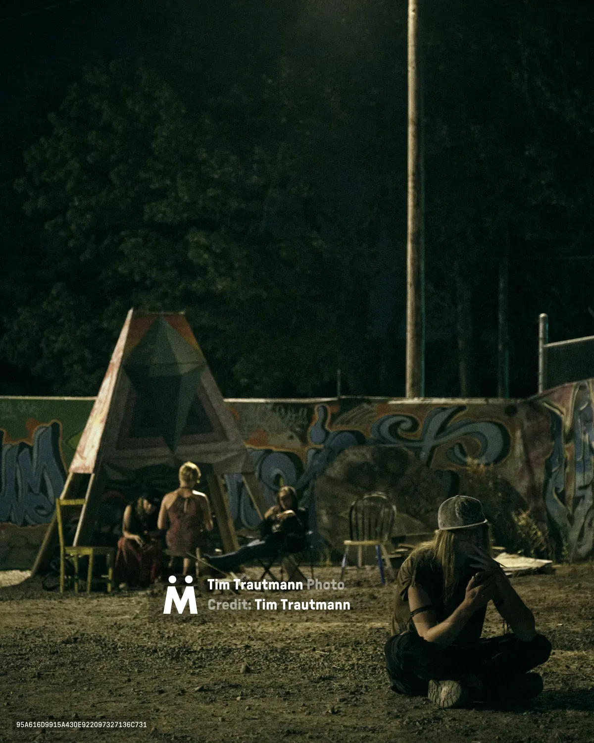 Under the amber glow of a single street lamp, members of a Portland art collective gather in their outdoor space on Southeast 76th Avenue. A man with long hair and baseball cap sits cross-legged in the foreground, while shadowy figures socialize among art installation structures and graffiti-adorned walls. The nocturnal scene captures the community spirit of Mount Tabor's underground creative collective, where artists connect in the darkness between day and dawn.