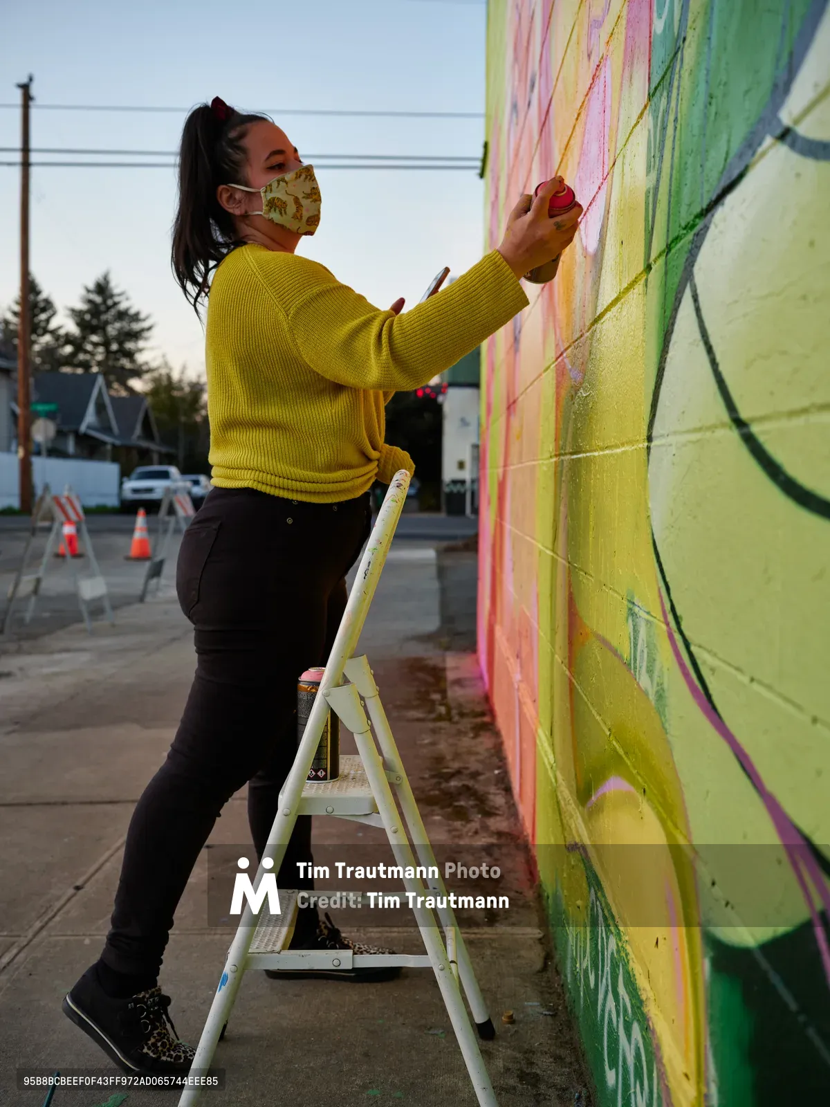 Multidisciplinary artist Villatose balances on a white stepladder, applying vivid streaks of pink and yellow paint to her towering wall mural 'Jardín Cubana' in Portland's Alberta Arts District. The artwork depicts three female figures rendered in flowing brushstrokes across the concrete canvas. Dressed in a mustard-yellow ribbed sweater and floral face mask, she works with focused determination as twilight descends over Northeast Alberta Street. The rainbow cascade of colors flows down behind her, while power lines and modest neighborhood buildings frame this moment of creative transformation in one of the city's most celebrated artistic corridors.