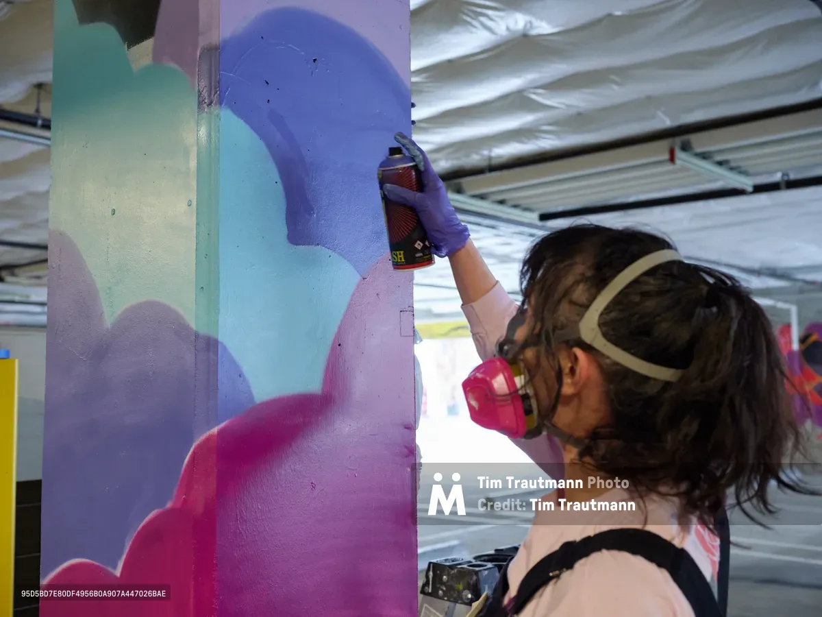 A young muralist wearing a protective respirator mask guides a spray paint canister across a concrete pillar, layering vibrant blues and magentas in flowing abstract forms. The industrial setting of Clay Creative's parking garage in Southeast Portland frames her focused work, with corrugated metal ceiling panels casting diffused light over the evolving artwork. Her concentrated posture and deliberate movements capture the meditative intensity of urban art creation in progress.
