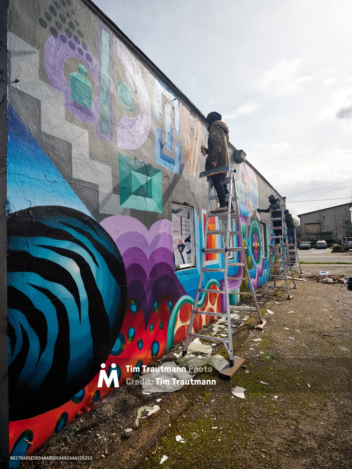 Two artists perched on aluminum ladders bring vibrant life to a concrete warehouse wall in Northeast Portland, their spray cans and brushes creating a kaleidoscope of geometric patterns and organic forms. The overcast Pacific Northwest sky provides soft, diffused lighting that makes the electric blues, magentas, and coral tones pop against the industrial backdrop. Paint cans and artistic debris scatter across the weathered asphalt below, evidence of the creative process unfolding on this urban canvas.