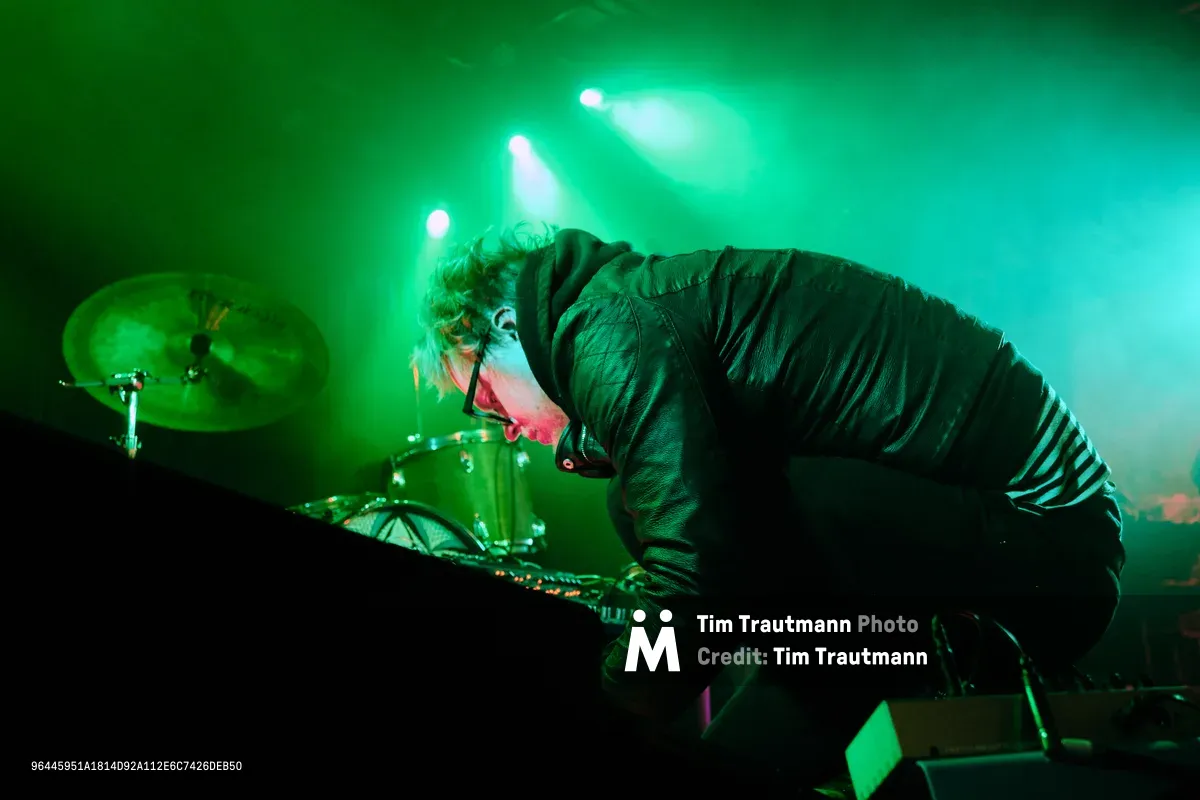 Electronic musician Eli Savage, performing as Ellipsism, hunches intensely over his equipment during a live set at RXFest 2019 in Portland's legendary Dante's venue. Bathed in ethereal green stage lighting that cuts through billowing fog, the artist's concentrated posture and wild curls create a dramatic silhouette against the luminous backdrop. The atmospheric haze and piercing spotlights transform the intimate club space into an otherworldly sonic landscape, with drum kit and electronic gear barely visible in the moody chiaroscuro.