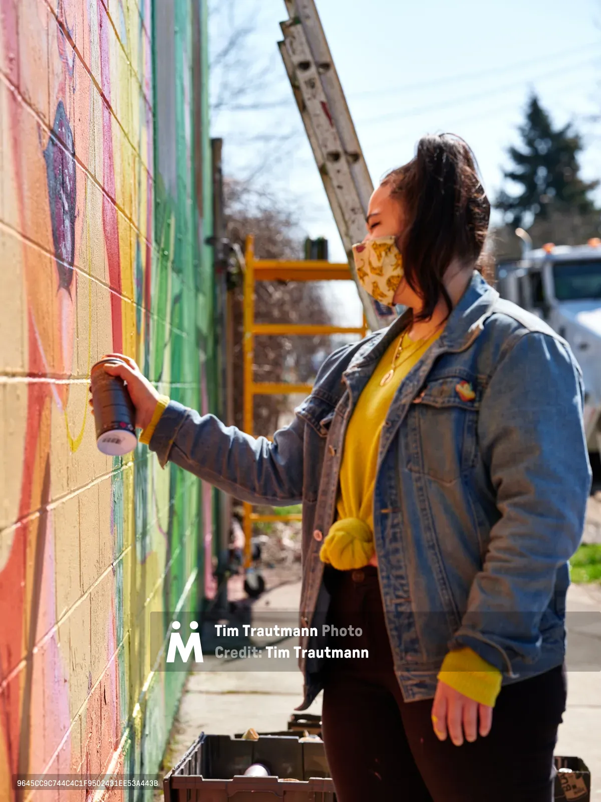 Multidisciplinary artist Vilatose works on Jardín Cubana, a mural depicting three female figures along Northeast Alberta Street in Portland's Alberta Arts District. Wearing a yellow bandana mask and denim jacket, the artist reaches toward a rainbow-hued wall featuring bold stripes of pink, yellow, and green paint. Golden afternoon light illuminates the urban scene, with construction equipment and bare winter trees visible in the background, capturing the ongoing transformation of this creative neighborhood.