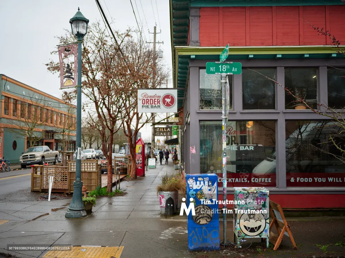 A slice of Portland's Alberta Street unfolds on an overcast day, where the Random Order Pie Bar's crimson storefront anchors a corner bustling with urban character. Street art adorns utility boxes while vintage lampposts and autumn-bare trees frame the sidewalk scene. The neighborhood's eclectic mix of colorful buildings and pedestrian activity captures the essence of this gentrified arts district.