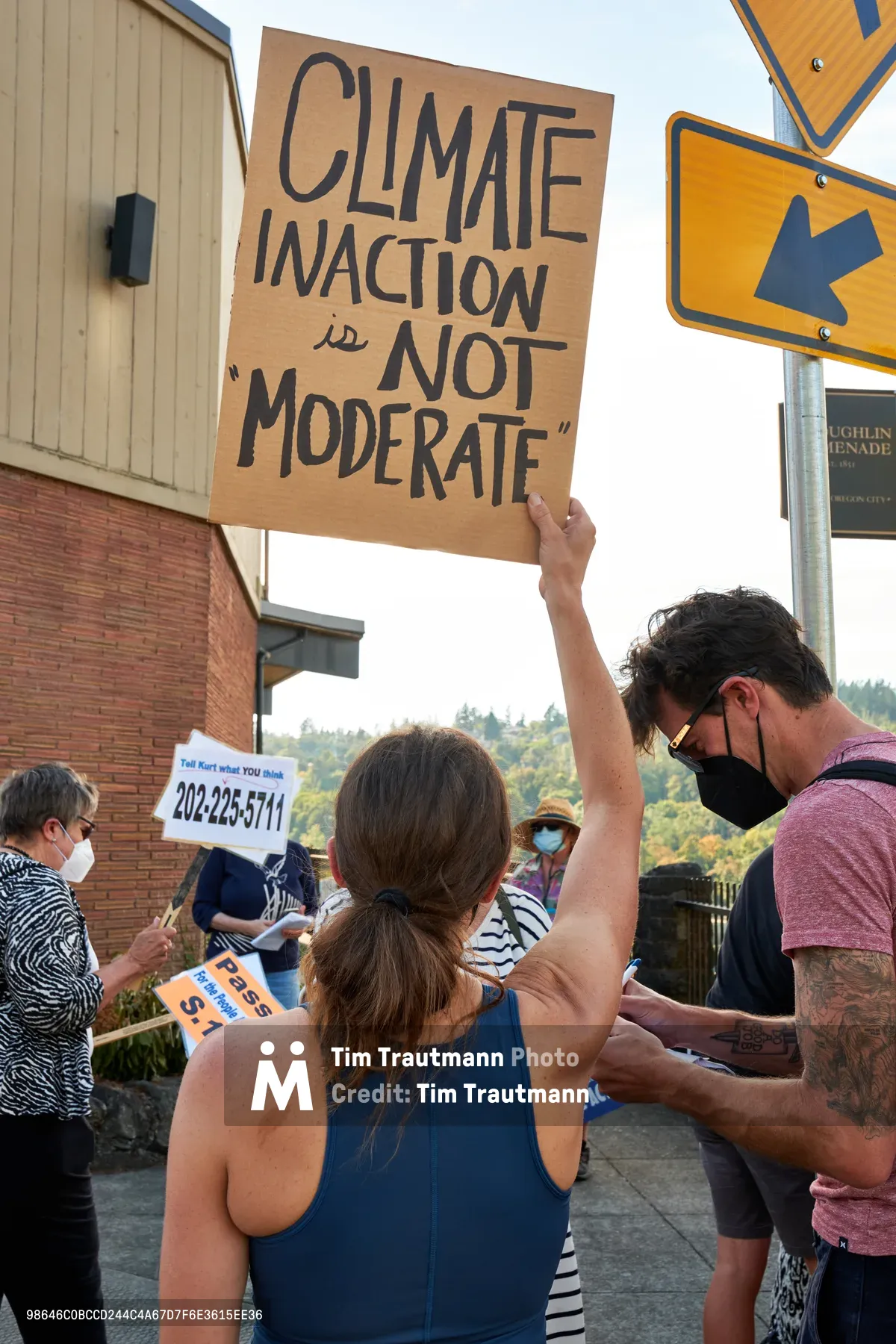 A woman in a blue tank top raises a handmade cardboard sign reading "CLIMATE INACTION IS NOT MODERATE" above her head during a demonstration outside Representative Kurt Schrader's office on McLoughlin Promenade. The protest unfolds under golden afternoon light, with masked participants holding various signs including phone numbers and political messages. Forested hills of Oregon City create a verdant backdrop behind brick municipal buildings and yellow directional street signs.