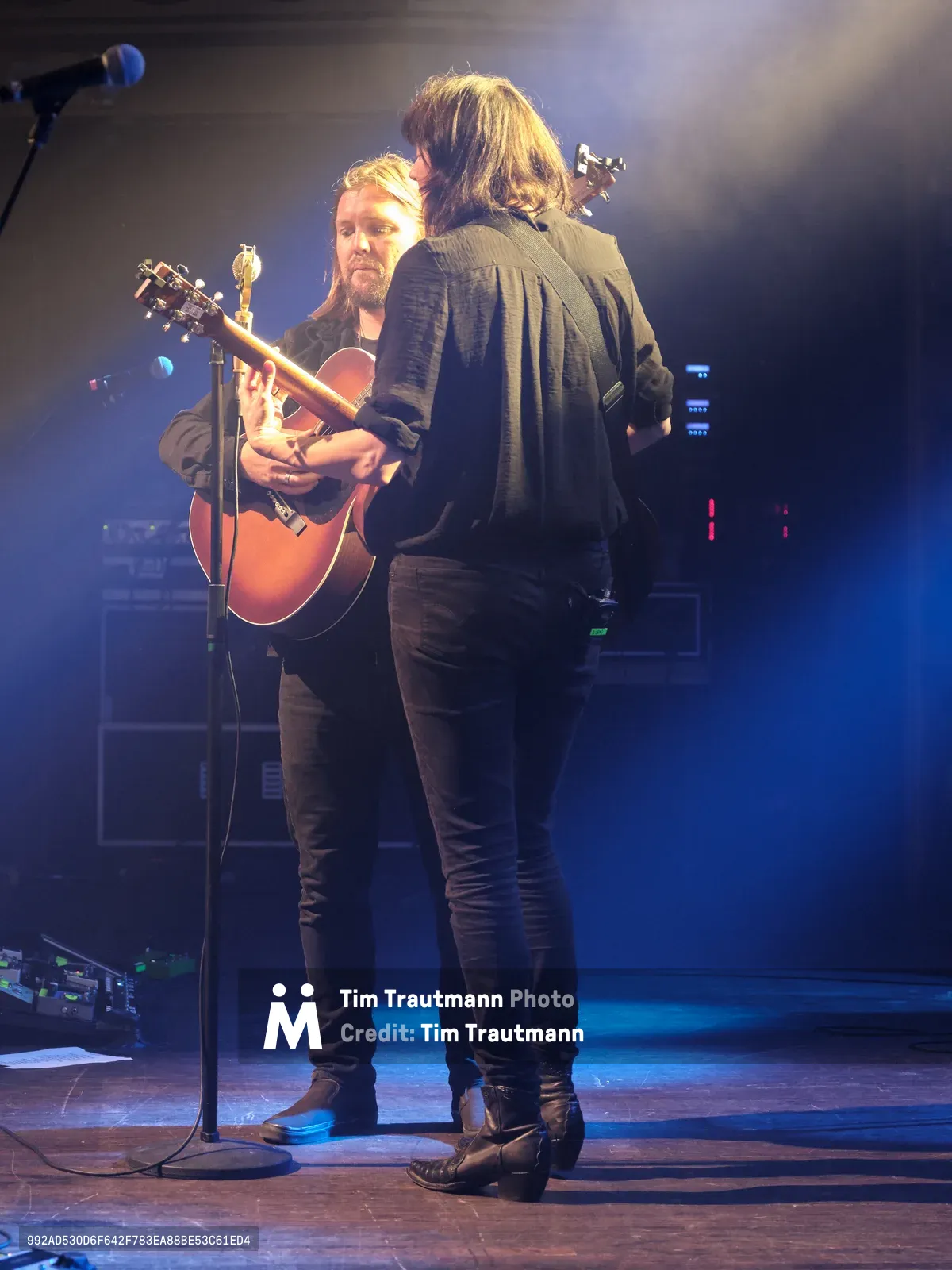 Emma Richardson and Russell Marsden of Band of Skulls share an intimate musical moment during their 10 Year Anniversary Tour at Webster Hall in Manhattan. Bathed in ethereal blue stage lighting with atmospheric haze, Richardson stands with her back to the audience while Marsden faces forward, cradling an acoustic guitar at the microphone. The dramatic lighting creates a moody, contemplative atmosphere that captures the emotional weight of their decade-long musical journey.