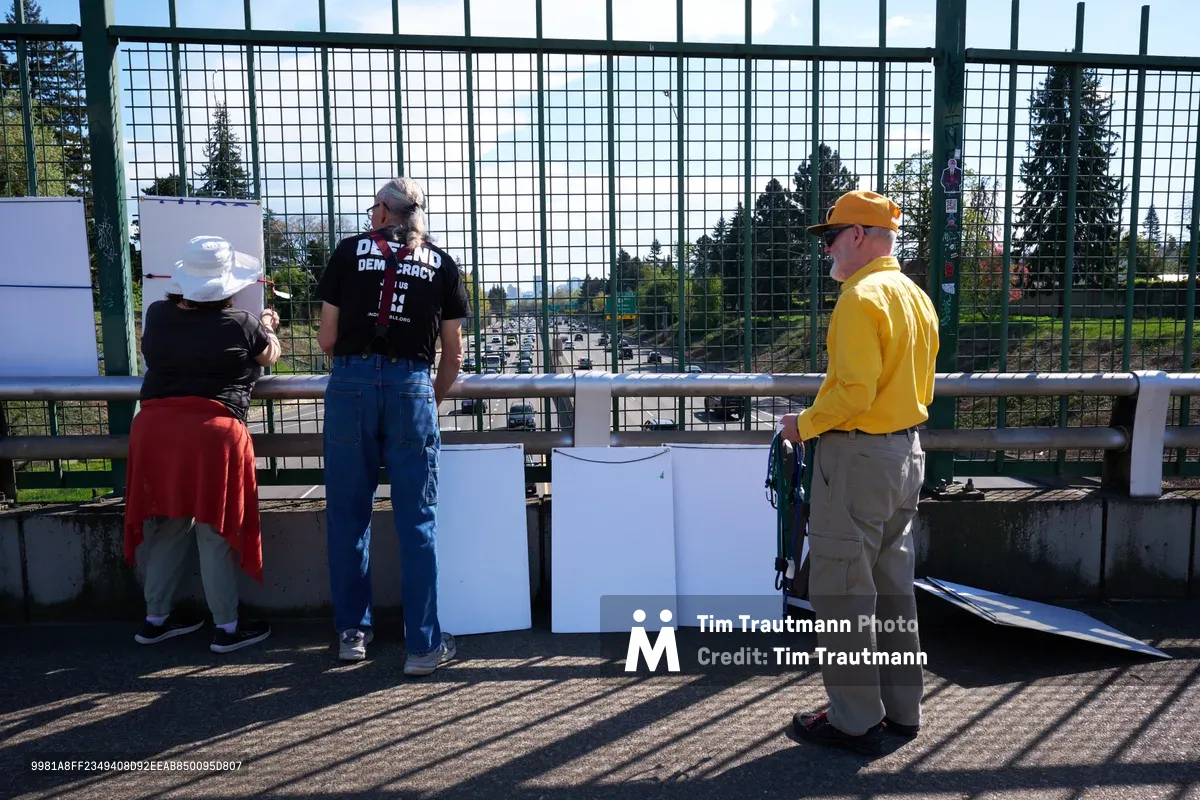 Three activists prepare for a coordinated banner drop on the North Skidmore Street overpass in Portland, Oregon, positioning themselves behind safety barriers above Interstate 5. Morning light filters through the protective mesh fencing as the participants—one in a white bucket hat and red shirt, another wearing a black 'Demand Democracy' t-shirt, and a third in bright yellow with an orange cap—organize their materials for the May 1st General Strike demonstration. The urban landscape of North Portland stretches beyond, with evergreen trees framing the highway corridor below.