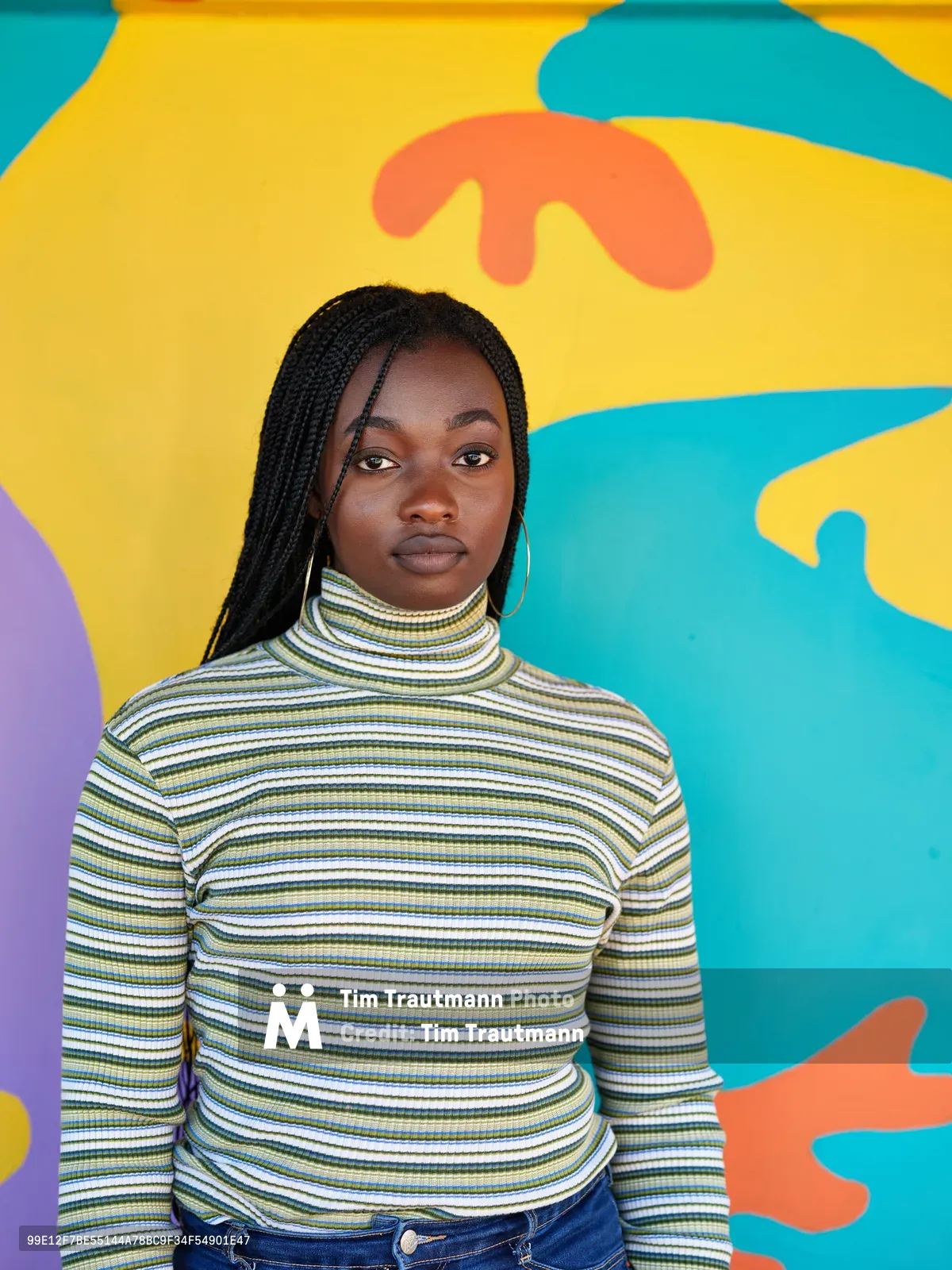 Naomi Likayi stands confidently before her vibrant street mural in Portland's Alberta district, wearing a striped turtleneck that echoes the rhythmic patterns of her artistic work. The kaleidoscopic backdrop bursts with flowing shapes in turquoise, golden yellow, coral, and purple, creating an energetic conversation between the artist and her creation. Soft, even lighting illuminates her direct gaze and the intricate box braids that frame her face, while the organic forms of the mural seem to dance around her figure.