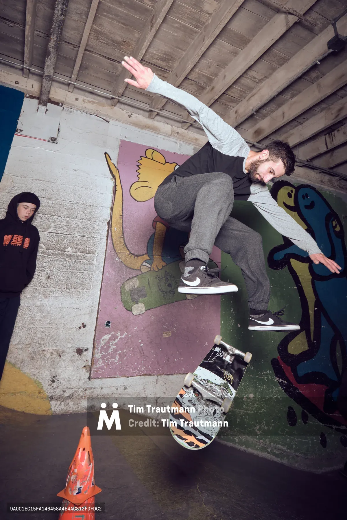 A bearded skateboarder executes a dynamic aerial maneuver in the concrete bowl at Commonwealth Skateboarding in Portland's Buckman neighborhood. The raw industrial space features exposed wooden beams overhead and colorful murals painted directly on the bowl's surfaces, including whimsical cartoon characters that contrast with the gritty concrete architecture. Another skater in a black hoodie observes from the sidelines as late afternoon light filters through the warehouse, creating dramatic shadows across the worn skating surfaces.