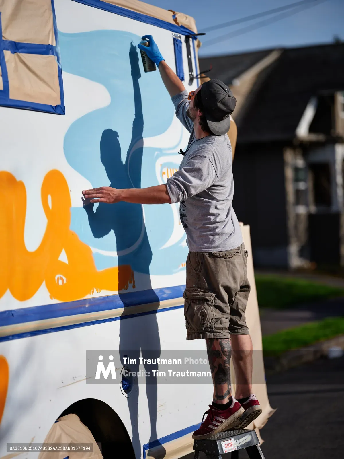 A tattooed muralist in a backwards cap and grey sweatshirt stretches upward with spray paint, adding vibrant orange and blue lettering to the side of a white food truck. Golden afternoon light illuminates his focused work as his shadow falls dramatically across the vehicle's surface. The residential Portland neighborhood setting creates an intimate backdrop for this moment of urban artistry in progress.