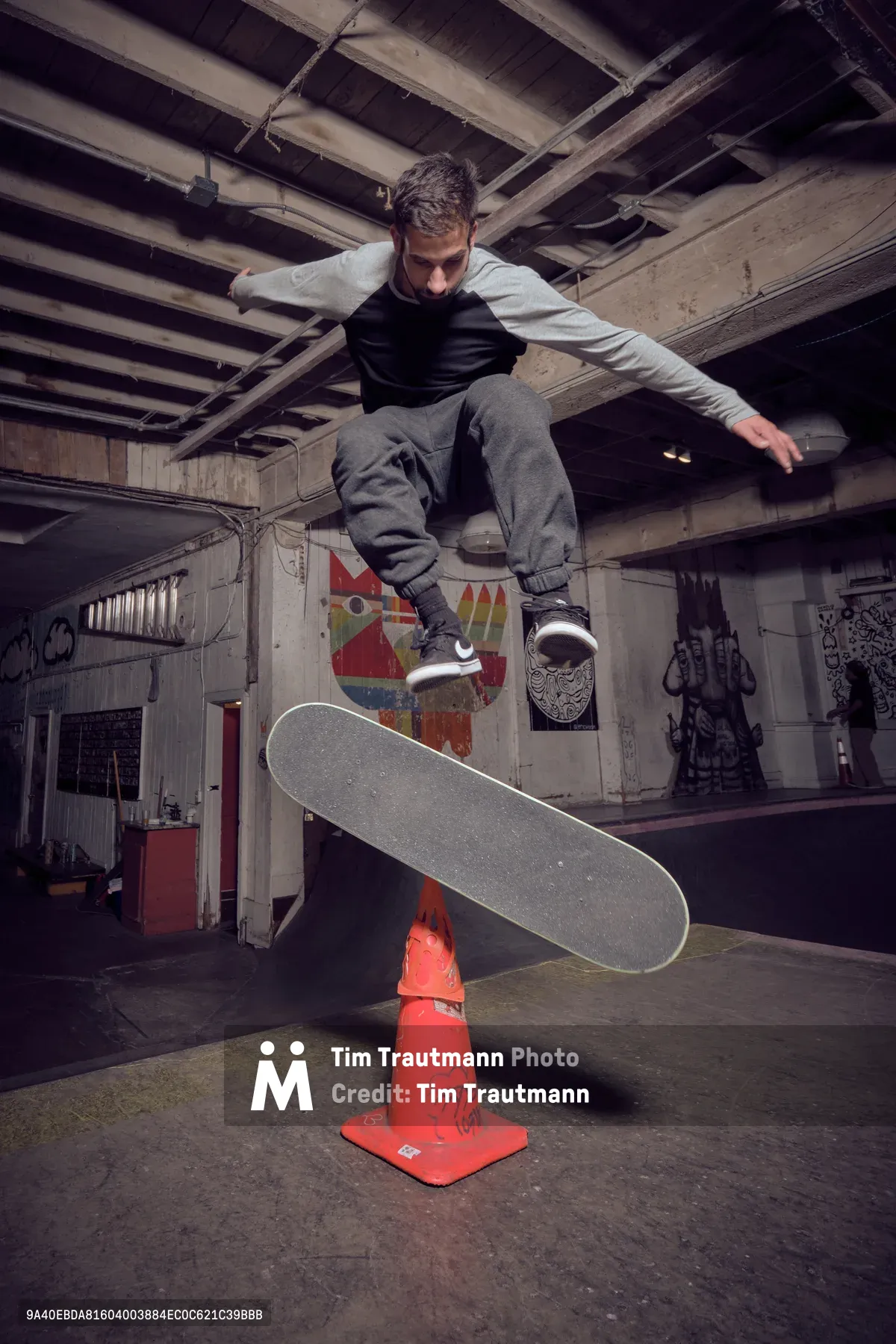 A young skateboarder executes a precise trick over an orange traffic cone in the raw, industrial confines of Commonwealth Skateboarding in Portland's Buckman neighborhood. Exposed wooden ceiling joists and graffiti-adorned concrete walls create a gritty backdrop, while dramatic lighting captures the athlete suspended mid-air above the improvised obstacle. The weathered concrete floor and urban decay aesthetic embody the authentic underground skateboarding culture that thrives in repurposed spaces.