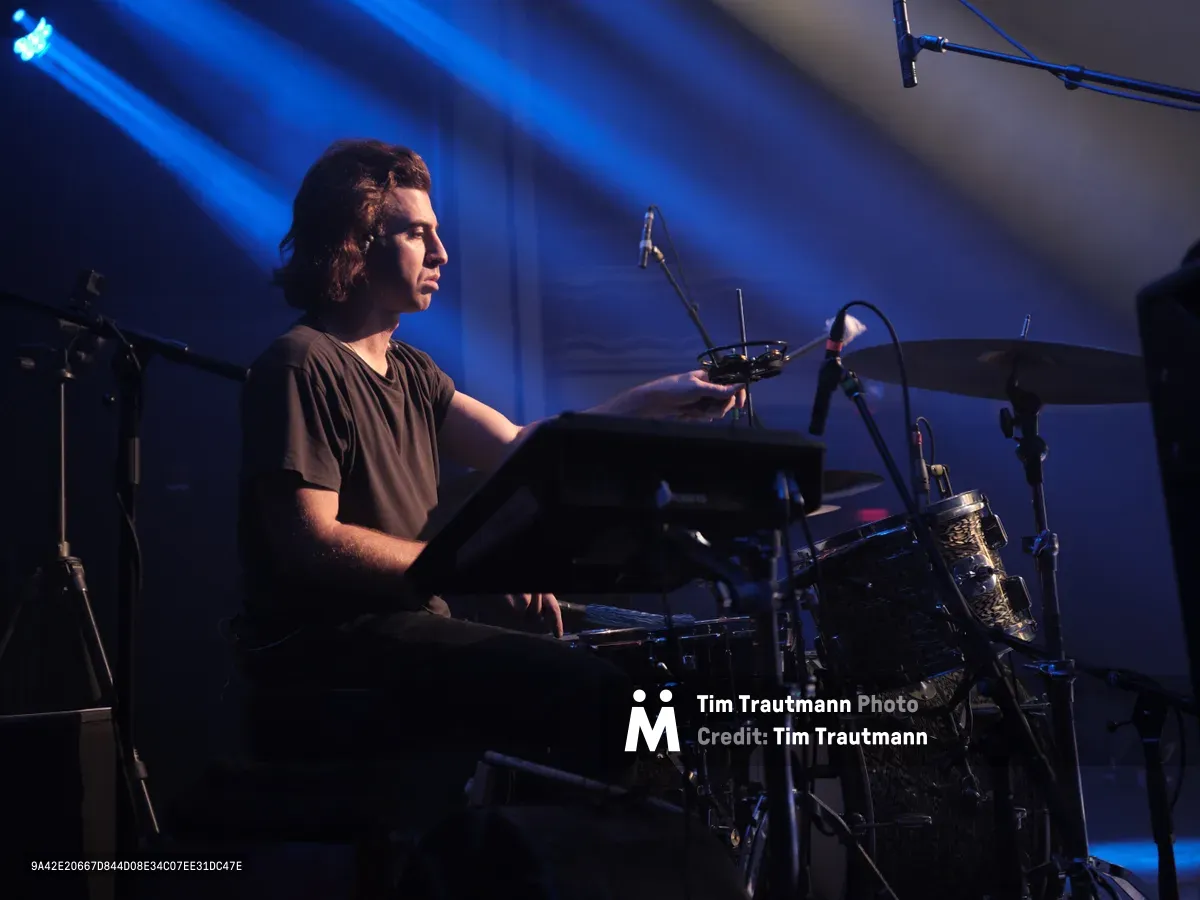 Drummer Julian Dorio of Band of Skulls sits poised behind his kit at Webster Hall during the band's 10 Year Anniversary Tour, bathed in ethereal blue stage lighting that cuts through the intimate venue's signature darkness. His focused expression and relaxed posture capture a moment of quiet intensity between songs, while overhead lighting rigs and microphone stands frame the scene. The atmospheric haze and dramatic chiaroscuro lighting transform the historic Manhattan music hall into a cathedral of sound.