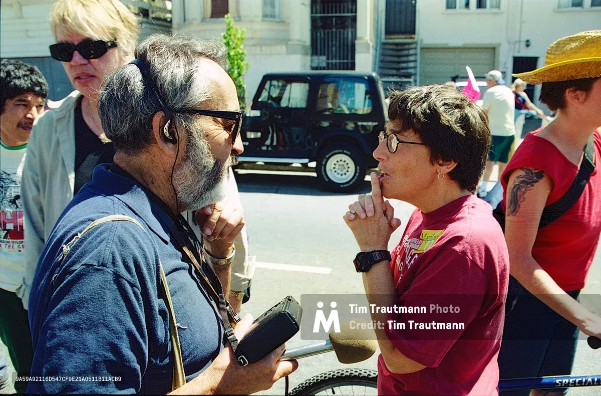 In the sun-drenched plaza at Bryant Street, a bearded journalist conducts an intimate street interview with a local activist during a Mission Anti-Displacement Coalition demonstration. The reporter leans forward intently, microphone extended, while his subject speaks passionately about gentrification's impact on San Francisco's historic Latino neighborhood. Behind them, fellow protesters gather in the harsh midday light, their presence adding urgency to this moment of grassroots media coverage.