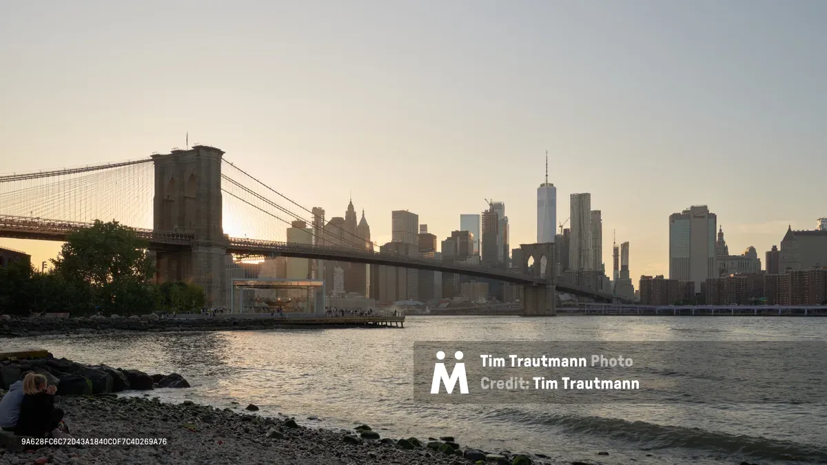 The iconic Brooklyn Bridge spans majestically across the East River as Manhattan's Financial District skyline rises behind it in warm twilight hues. Shot from the rocky shoreline of DUMBO's Brooklyn Bridge Park, the scene captures the serene transition from day to night with gentle waves lapping against moss-covered stones in the foreground. A lone figure sits contemplatively on the waterfront rocks, dwarfed by the architectural grandeur of both the historic suspension bridge and the gleaming towers of One World Trade Center beyond.