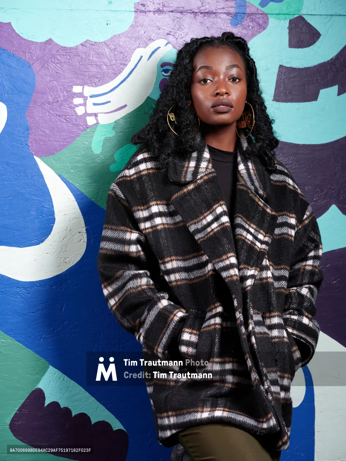 Artist Naomi Likayi stands confidently against her vibrant mural work at Portland's World Trade Center, her dark curls framing a direct gaze toward the camera. She wears an oversized plaid coat in earth tones over a black top, complemented by gold hoop earrings that catch the natural light. The abstract mural behind her bursts with swirling forms in blues, purples, and mint green, commissioned by the Portland Street Art Alliance for the boarded-up building. The composition creates a striking contrast between the artist's grounded presence and the dynamic energy of her colorful creation.