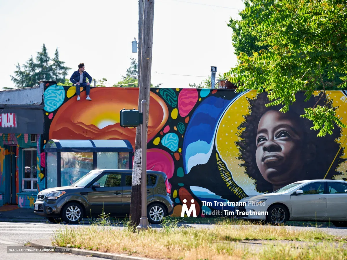 Muralist Travis Fields sits triumphantly atop the completed rooftop of a building adorned with his vibrant portrait of poet Amanda Gorman in Portland's Alberta district. The expansive mural features Gorman's face surrounded by a golden halo and kaleidoscopic patterns of turquoise, magenta, and orange that cascade across the building's facade. Two parked cars rest beneath the artwork while lush green foliage frames the scene under an overcast Pacific Northwest sky.